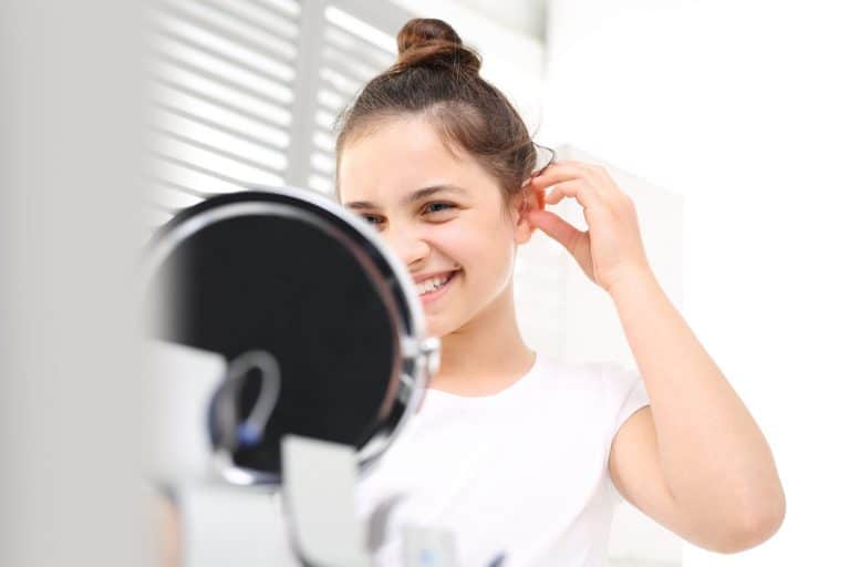 A woman putting on a hearing aid while smiling at a mirror.