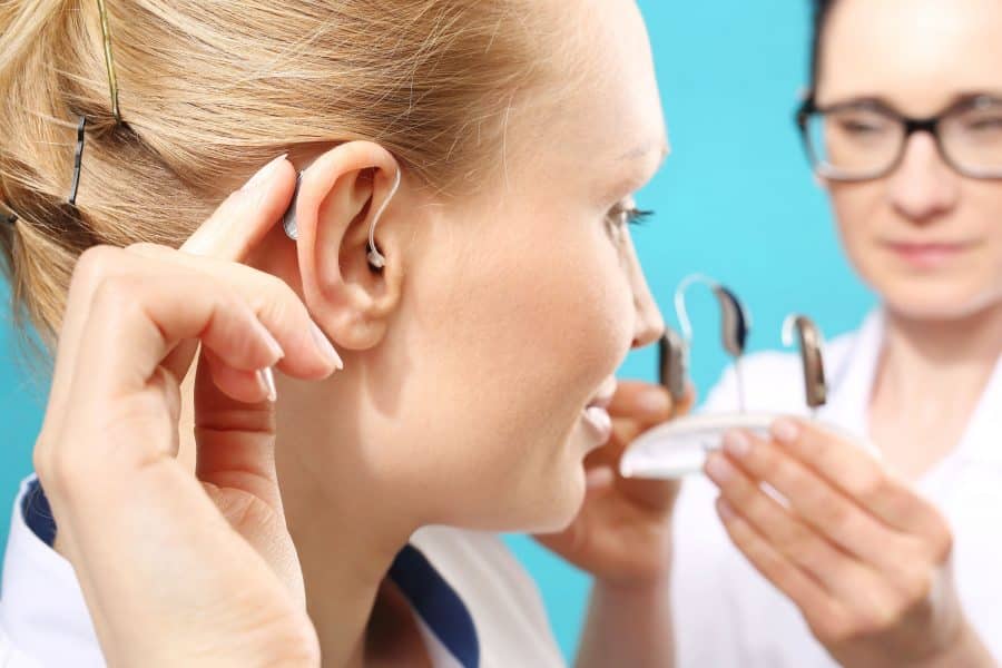 Close up of a woman’s ear with a hearing aid while a specialist holds hearing aids behind her.